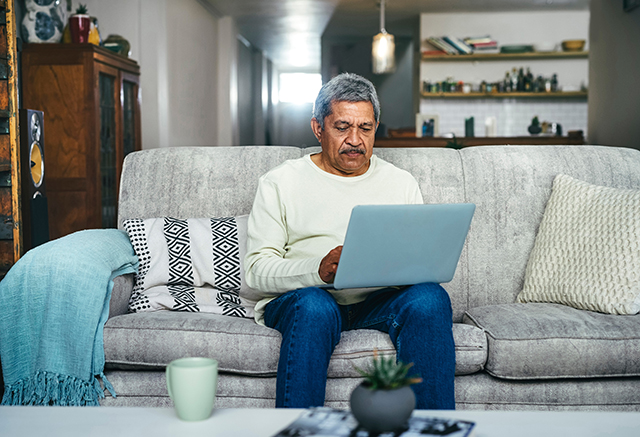 An aged man sits on a gray couch. He's wearing a white long-sleeved shirt and jeans. He's browsing the internet on his computer.
