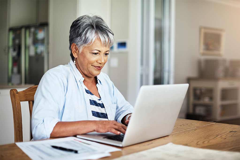 An older woman sits at her kitchen table, searching for something on her computer.