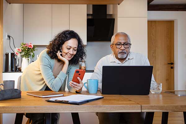 A couple stands around their dining room table using their phone and computer to schedule an appointment.
