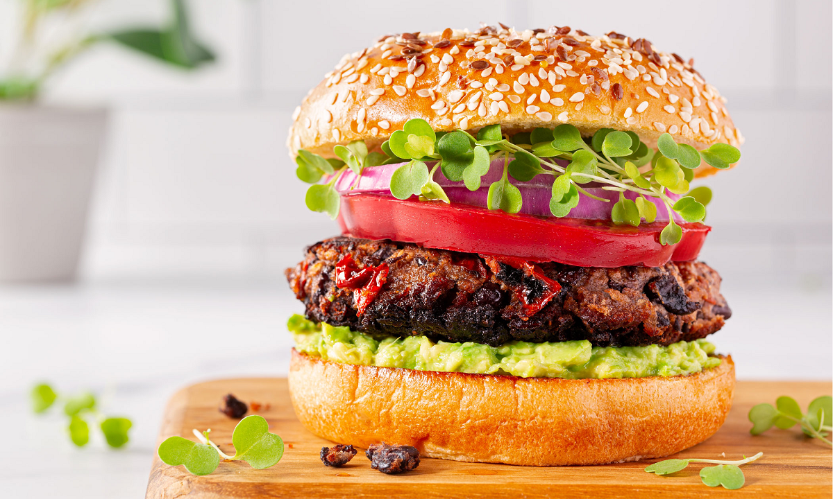 Black bean burger in a sesame seed bun, on top of a wood cutting board layered sprouts, red onion, tomato and avocado mash.