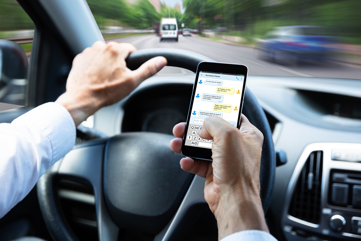 Person with one hand on a car steering wheel while their other hand sends a text message.