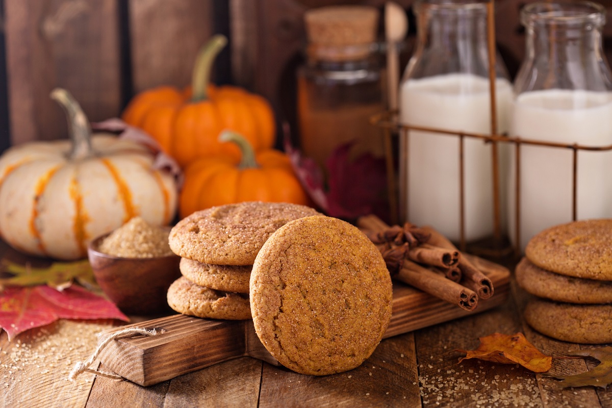 Brown cookies and whole cinnamon sticks on wooden cutting board in front of three small pumpkins and glass milk cartons.