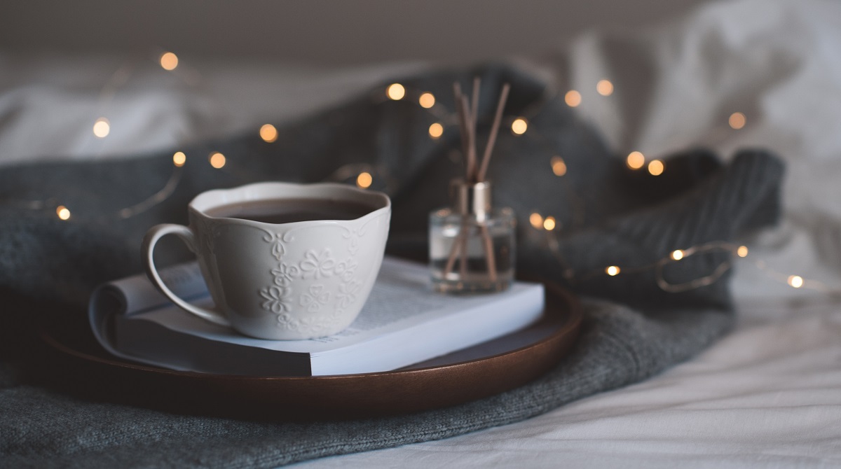 White cup of warm team on top of an open book and surrounded by twinkling lights and difuser.