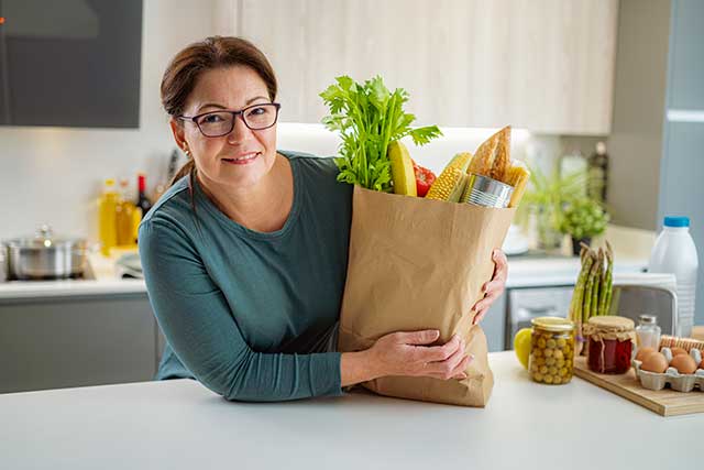 A middle-aged woman smiles as she leans over a bag of fresh groceries in her kitchen, including celery and bread inside